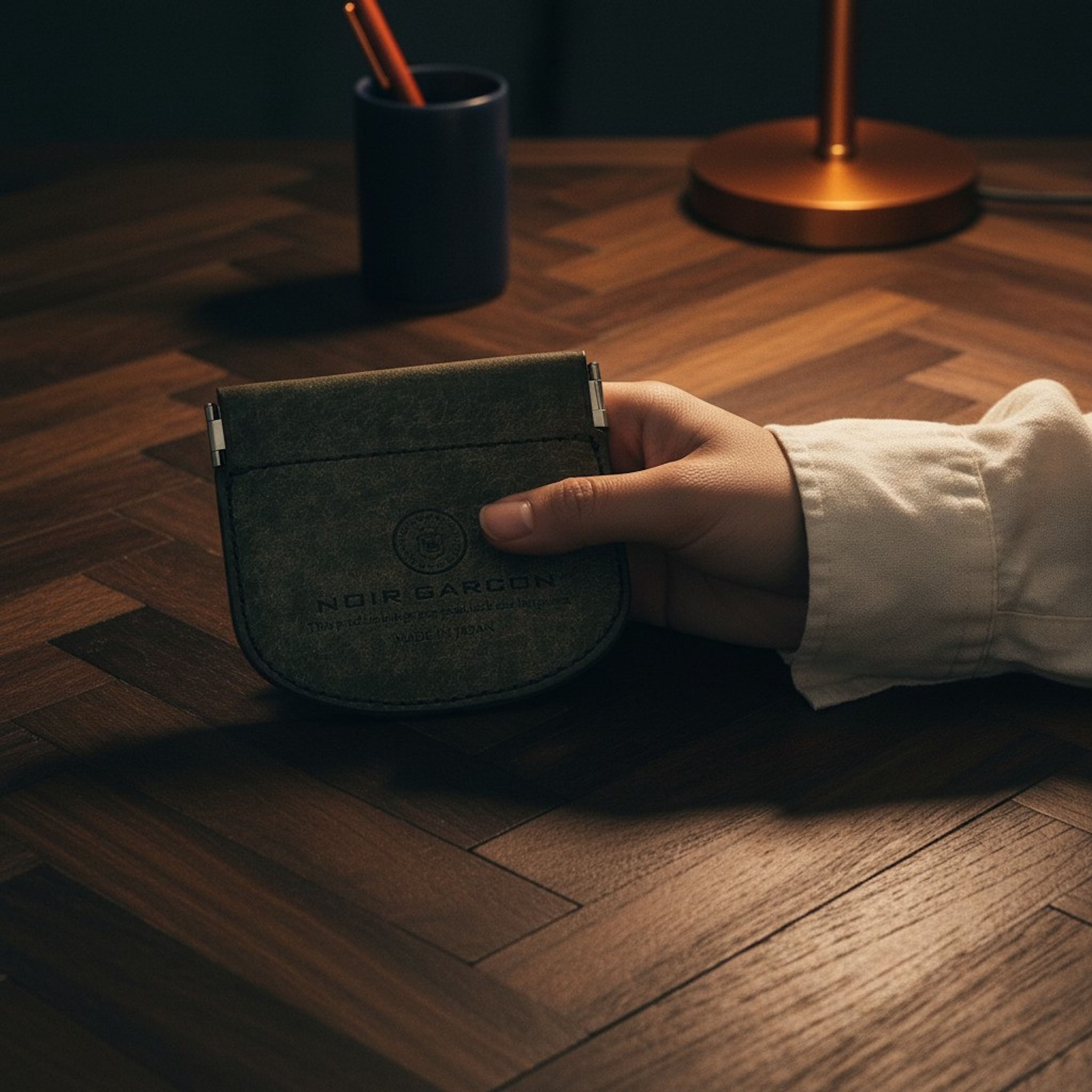 A hand holding a dark green velvet card holder with the brand name 'MOIR GARNIER' printed on the front, set on a dark wood herringbone table with a lamp and cup in the background.