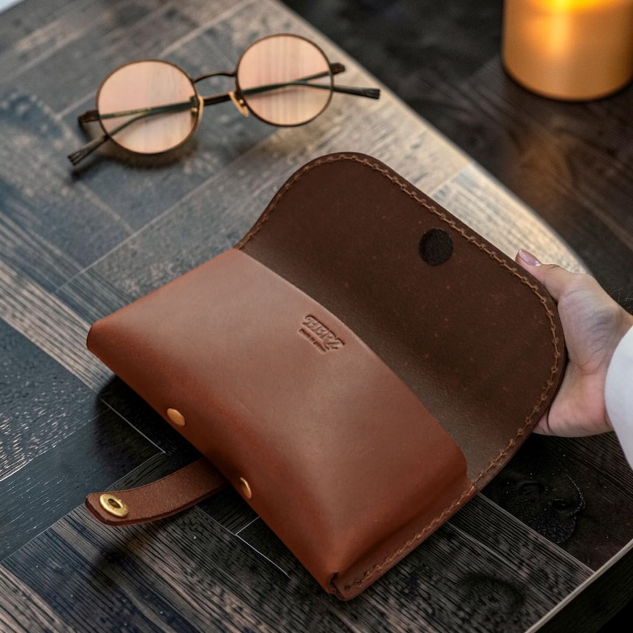 Brown leather eyewear bag held by a hand on a wooden surface with glasses in the background