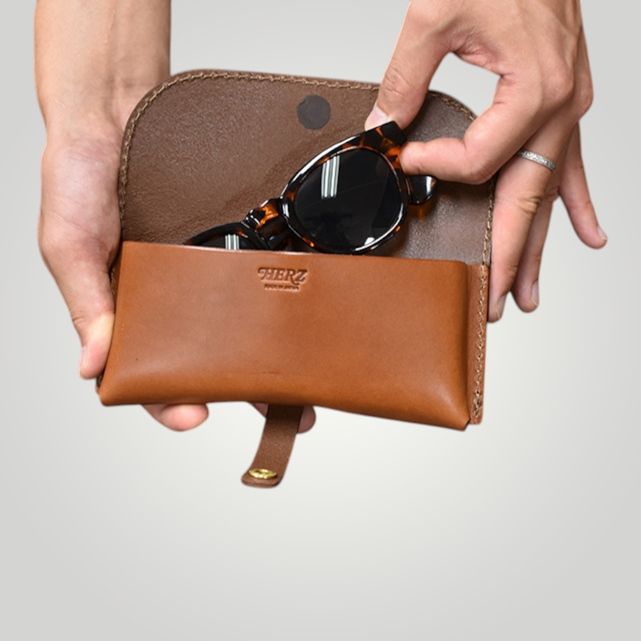 Brown leather eyewear bag with sunglasses held by a hand on a light gray background