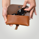 Brown leather eyewear bag with sunglasses held by a hand on a light gray background