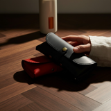 Hand holding a black leather eyewear bag with a red interior on a wooden floor.