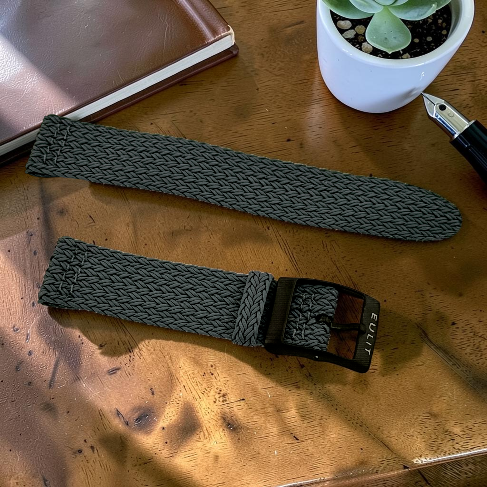 Two dark green woven watch straps on a wooden surface with a plant and notebook in the background.