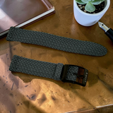 Two dark green woven watch straps on a wooden surface with a plant and notebook in the background.