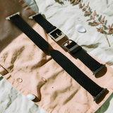 Black woven watch strap on a textured surface with dried leaves.