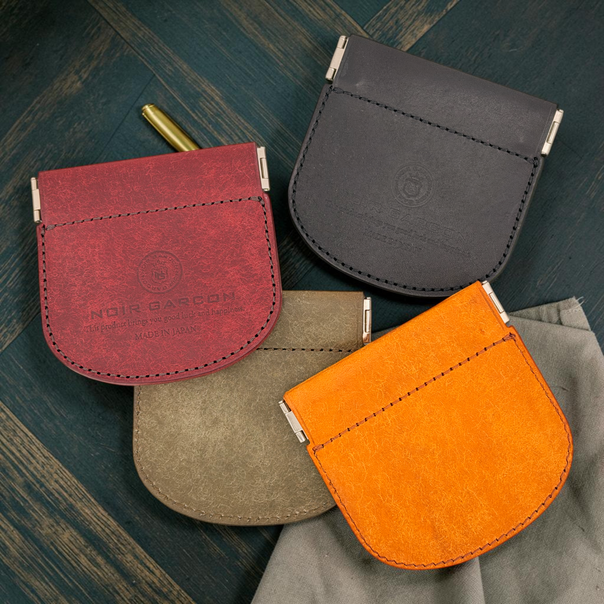 Four small leather card pouches in red, dark gray, beige, and orange, arranged on a dark wood surface with a gray cloth and a gold pen as props. The pouches show debossed branding, including Noir Garcon.