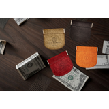Four small leather card holder wallets in gold, dark brown, red, and orange, arranged on a dark wood surface with stacks of cash around them. The wallets are embossed with the Nour Bamson logo.