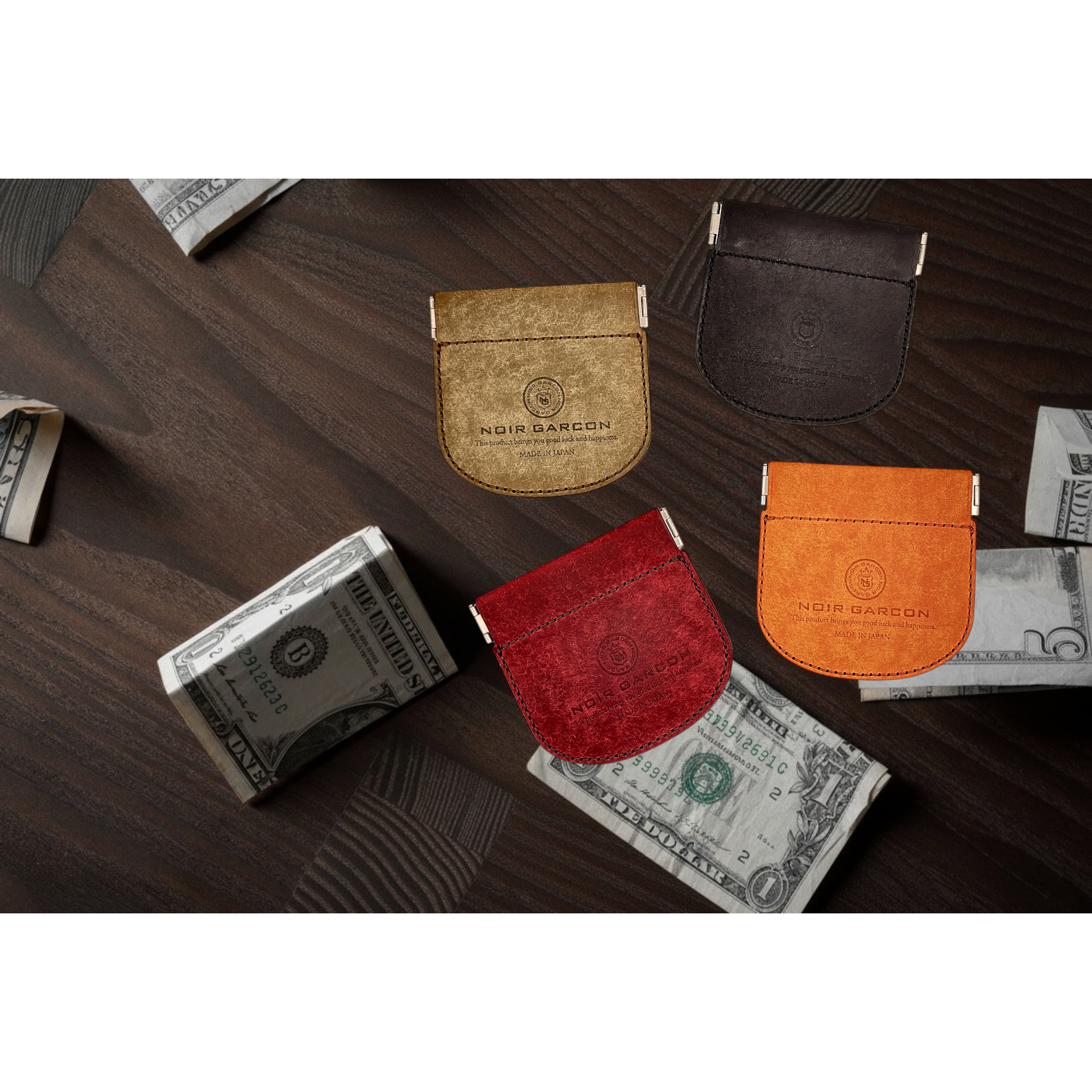 Four small leather card holder wallets in gold, dark brown, red, and orange, arranged on a dark wood surface with stacks of cash around them. The wallets are embossed with the Nour Bamson logo.