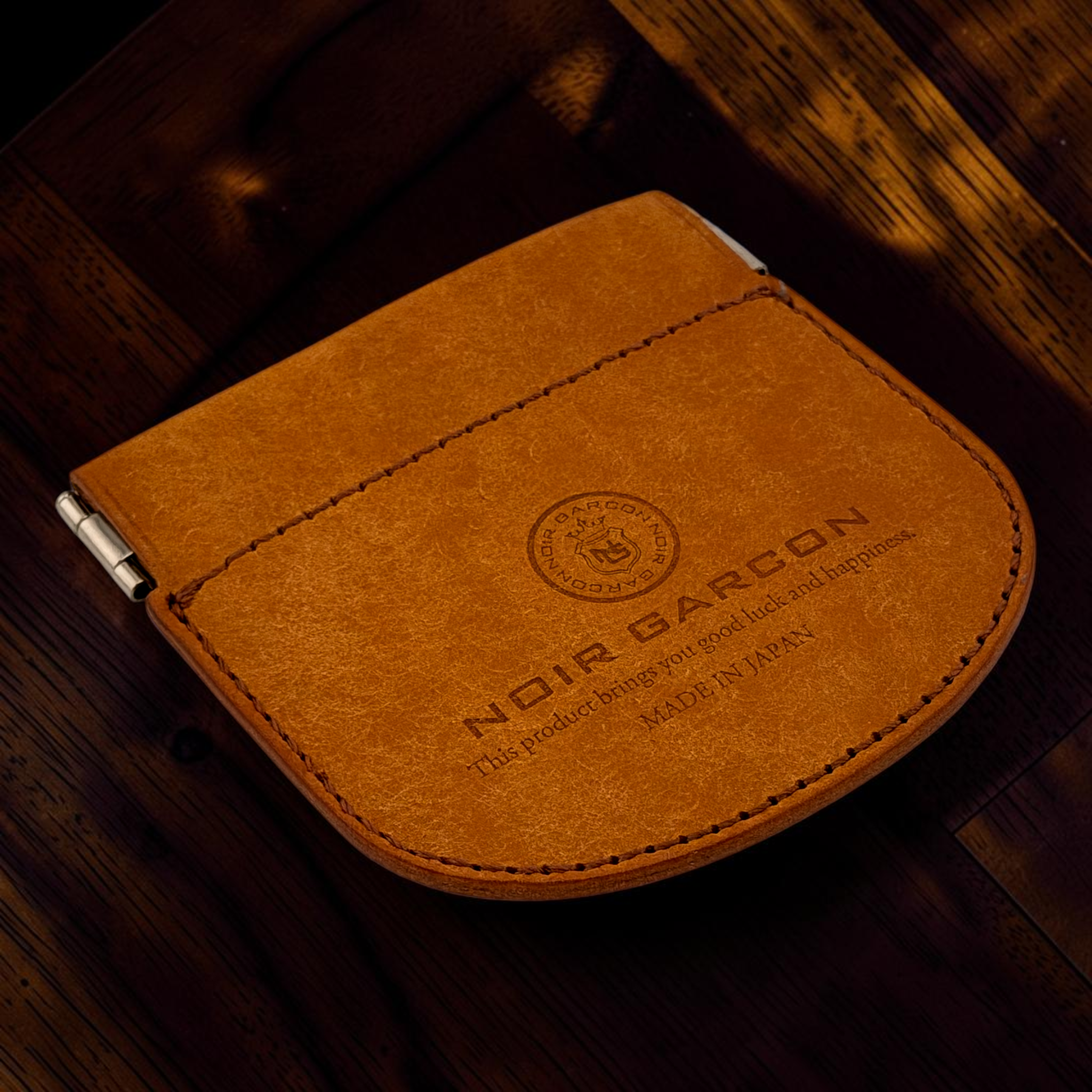 A close-up of a brown leather card holder or wallet with visible stitching, a metal clip on the side, and embossed branding reading 'NOIR BARCON' on a dark wooden surface.