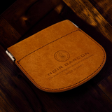 A close-up of a brown leather card holder or wallet with visible stitching, a metal clip on the side, and embossed branding reading 'NOIR BARCON' on a dark wooden surface.