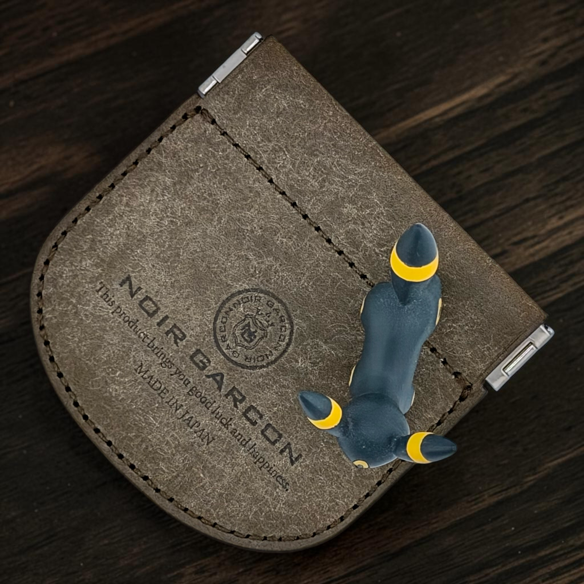 A brown textured leather card holder wallet with silver metal clips, featuring an embossed circular logo and the text 'NOIR BARCON'. A small dark blue fox figurine with yellow accents is placed on top of the wallet, which is photographed on a dark wood background.