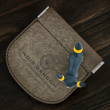 A brown textured leather card holder wallet with silver metal clips, featuring an embossed circular logo and the text 'NOIR BARCON'. A small dark blue fox figurine with yellow accents is placed on top of the wallet, which is photographed on a dark wood background.