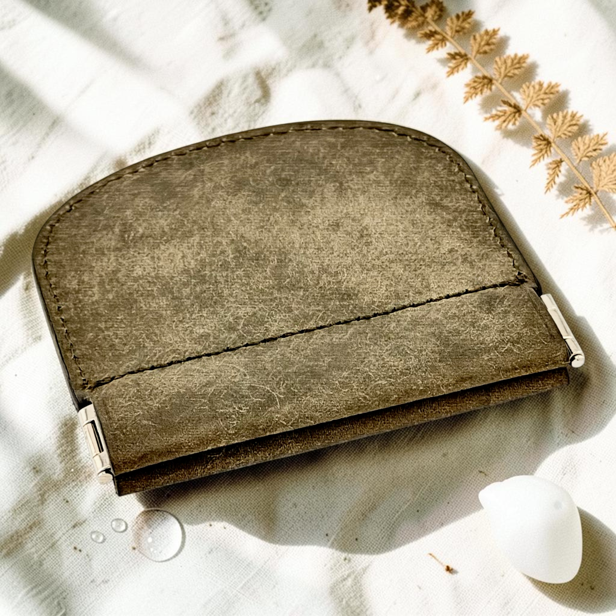 A small leather card holder or wallet in a muted taupe-brown color, styled on a textured white fabric with dried botanicals and a white stone.