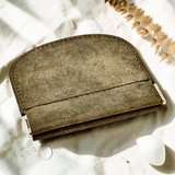A small leather card holder or wallet in a muted taupe-brown color, styled on a textured white fabric with dried botanicals and a white stone.