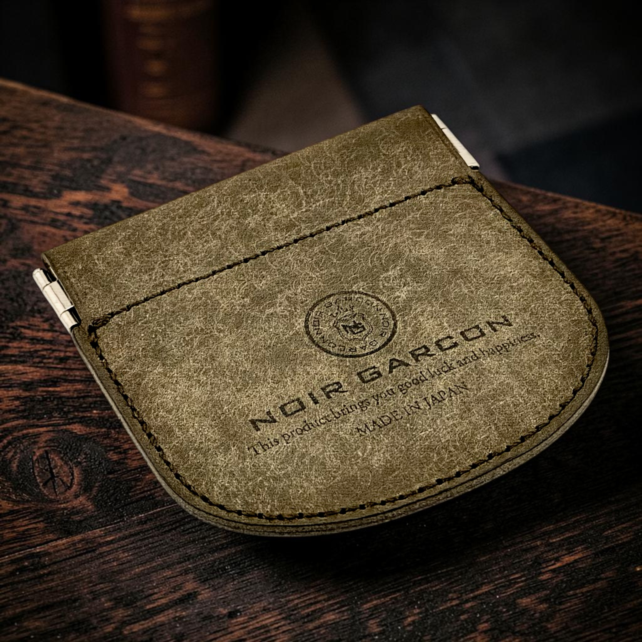 A close-up of a textured olive green leather money clip wallet with 'NOIR GARCON' branding, resting on a dark wooden surface.