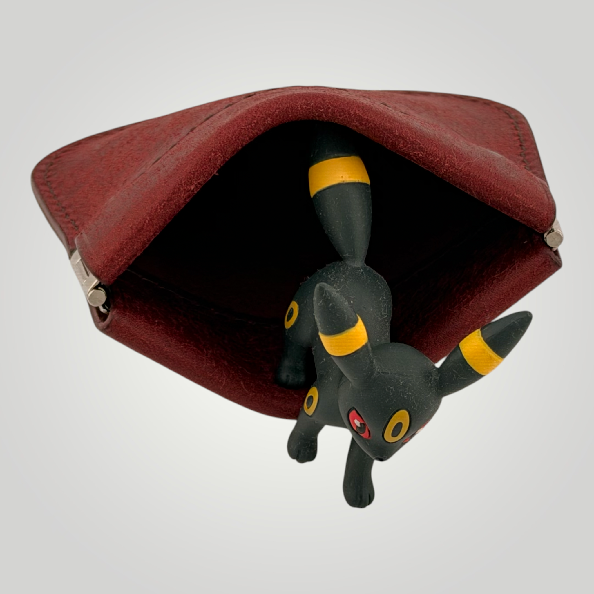 A small black toy figure with yellow bands and a red eye detail, partially tucked inside an open dark red pouch against a light gray background.