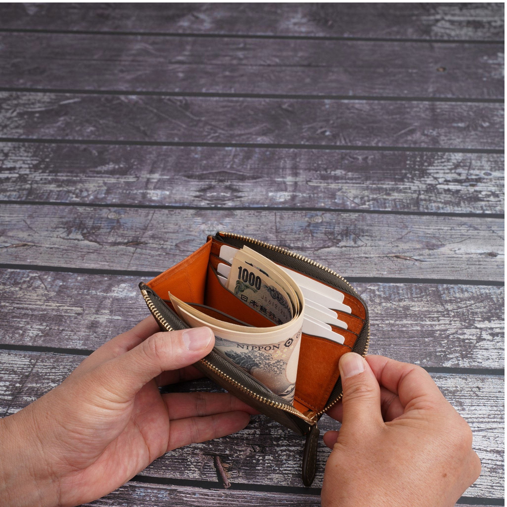 A small brown leather wallet with gold-tone zippers and an orange interior, shown open with cash and cards inside. Two hands are holding the wallet over a gray wood plank surface.